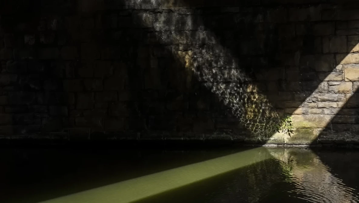 Sunlight streaming through a narrow passageway between dark stone walls and still green water below.