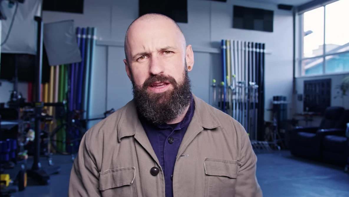 Bearded man with shaved head wearing tan work shirt in a modern studio with blue flooring and equipment racks.