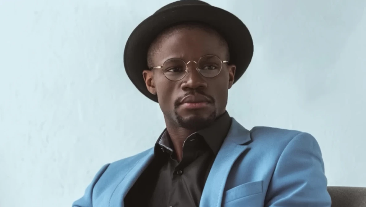 Man wearing blue blazer, black shirt, and glasses looks directly at camera with neutral expression.