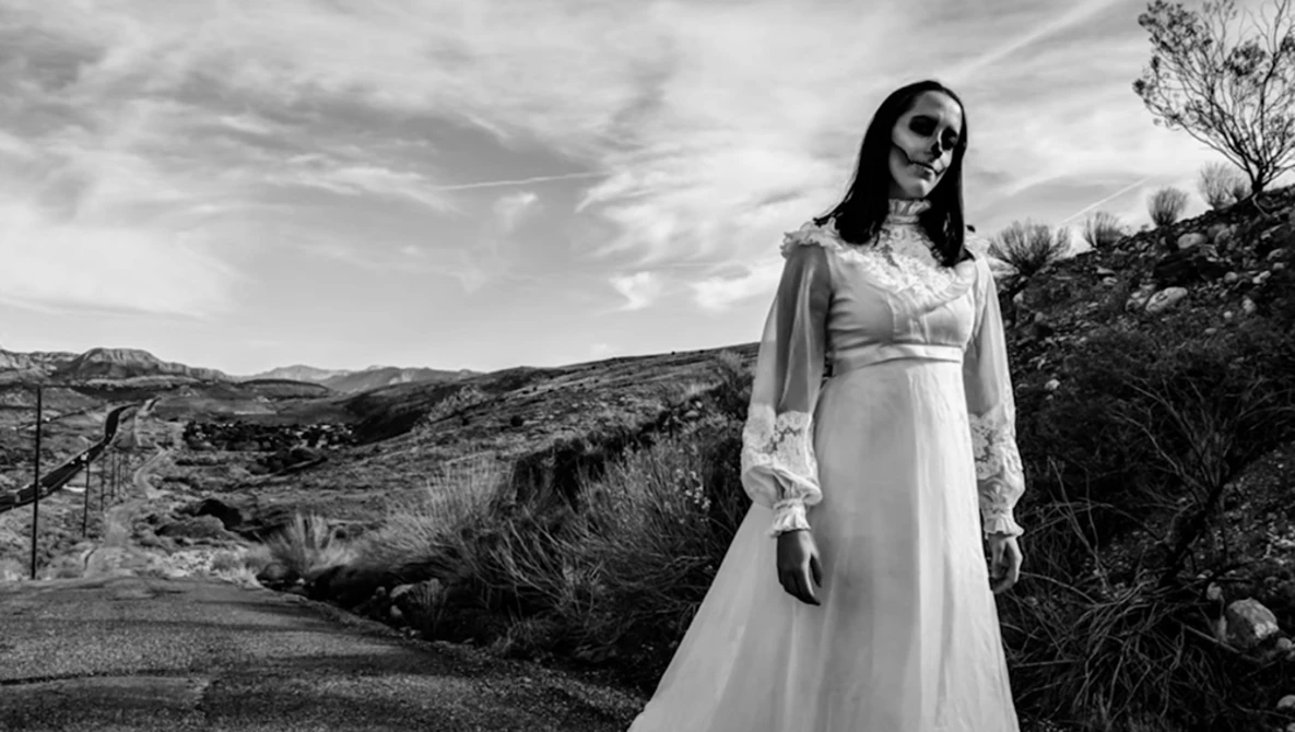 Person with skull face makeup wearing white flowing dress in desert landscape.