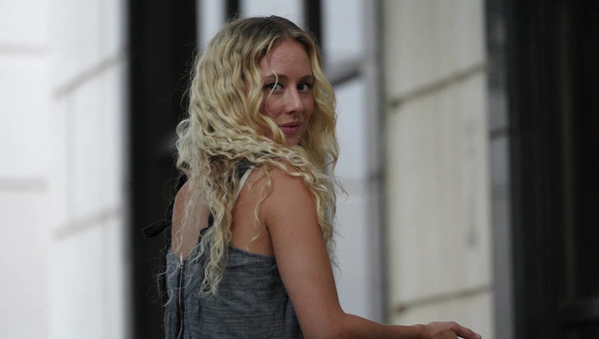 Woman with long blonde wavy hair wearing a gray sleeveless top, looking over her shoulder in an indoor hallway.
