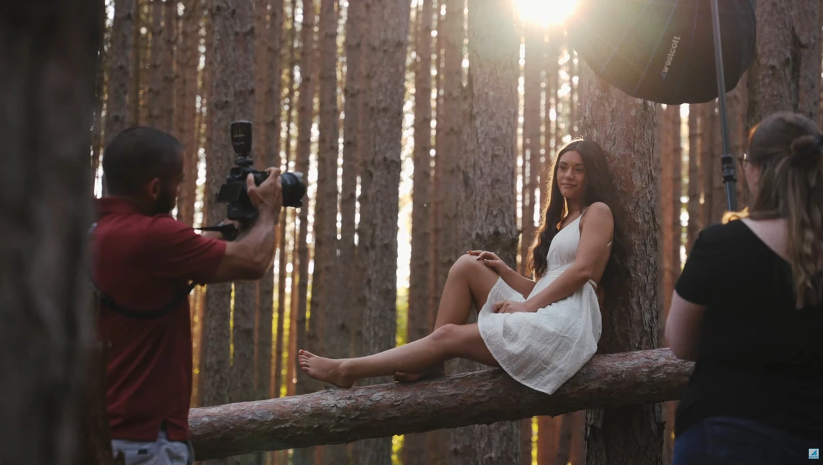 Female model sitting on a fallen log in a forest while being photographed by a crew member using professional lighting equipment.