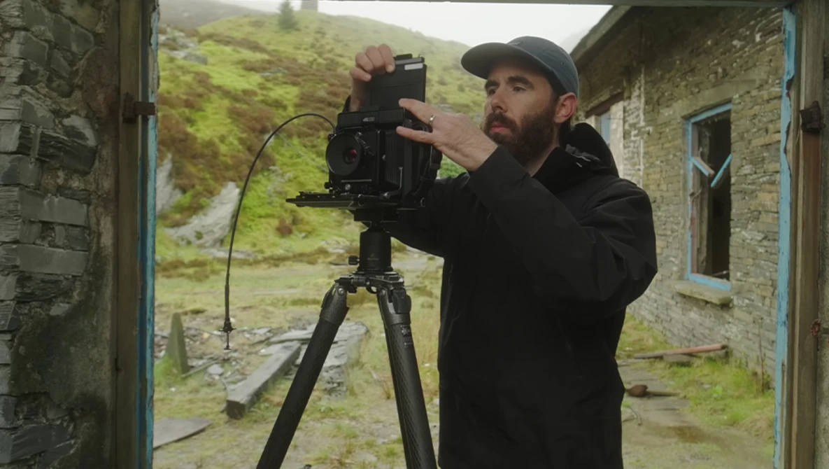 Filmmaker operating a cinema camera mounted on a tripod inside a stone building doorway.