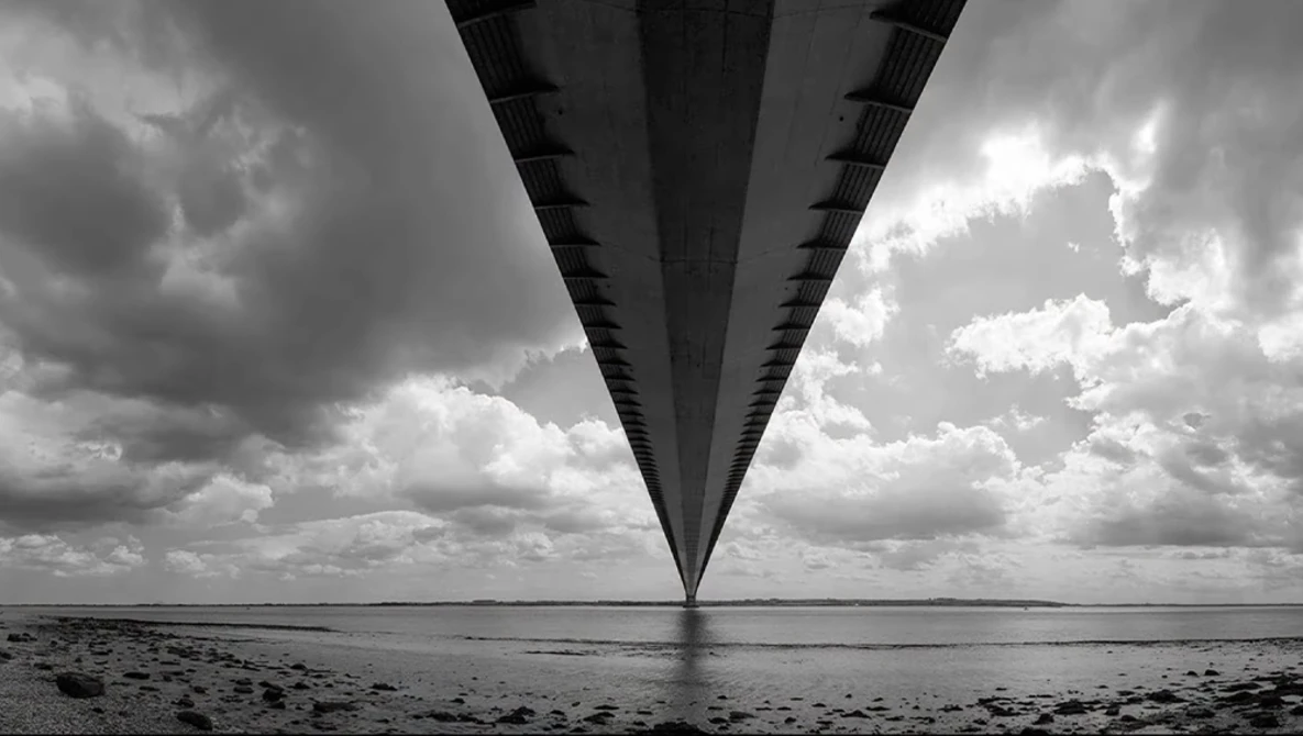 Long bridge viewed from underneath, stretching toward horizon with dramatic cloudy sky.