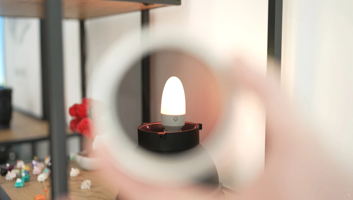 LED candle with glowing white flame inside dark holder, viewed through blurred circular glass or lens.