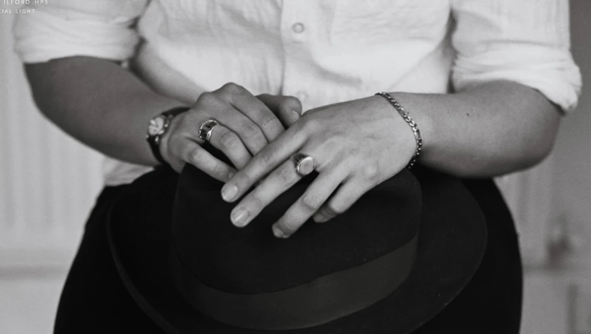 Close-up of two people's hands with wedding rings resting on a dark hat, shot in black and white.