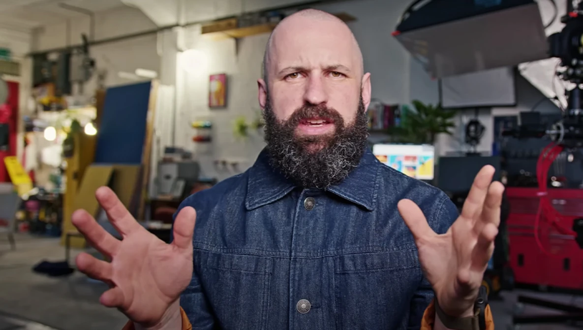 Bald man with full beard wearing denim shirt gesturing while speaking in a workshop studio.