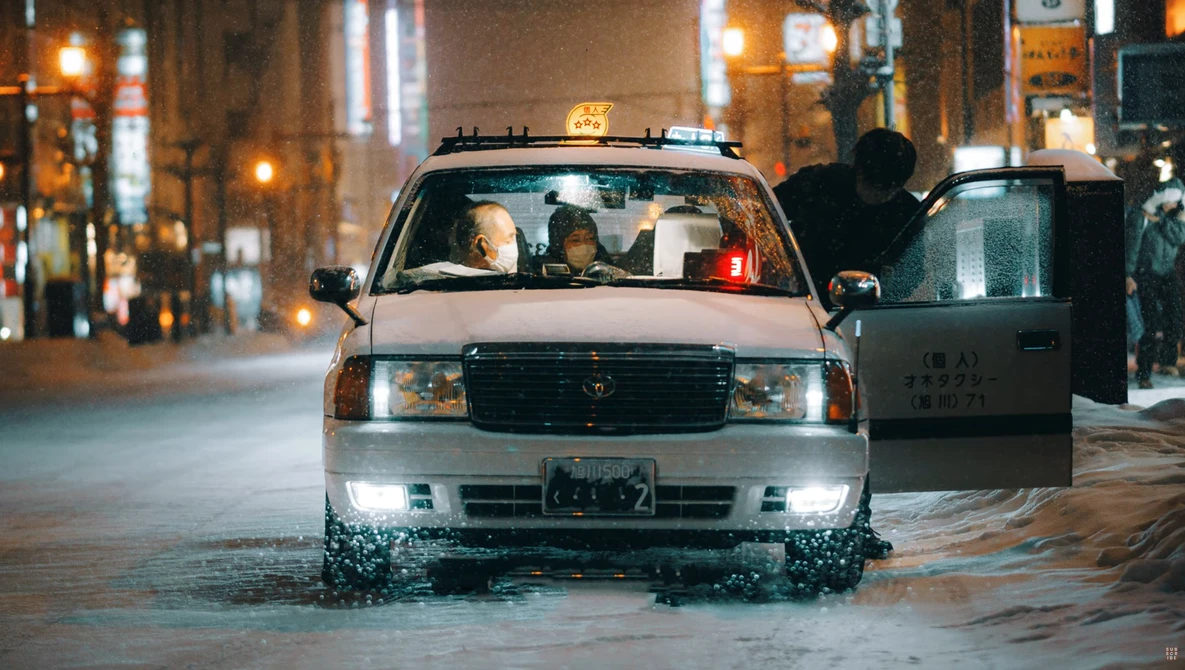Taxi cab driving through flooded urban street at night with city lights in background.