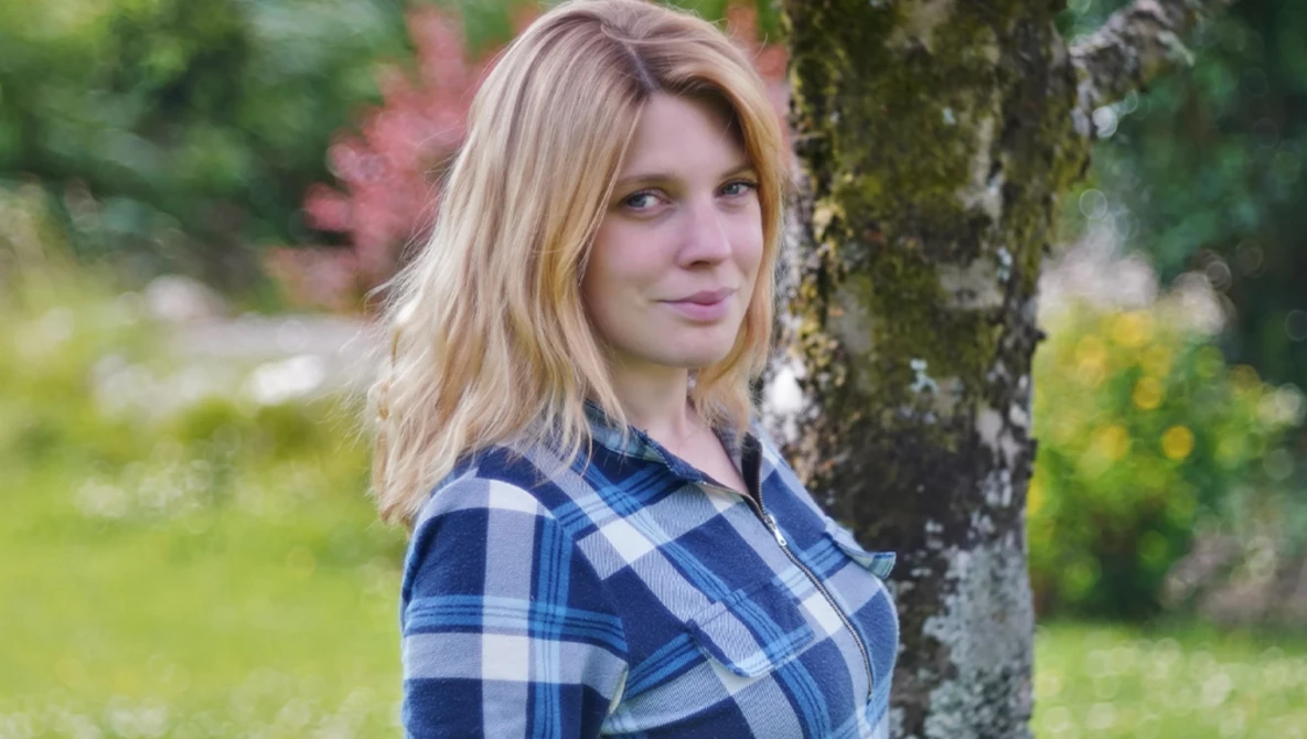 Woman in blue plaid shirt leaning against a moss-covered tree trunk in a garden setting.