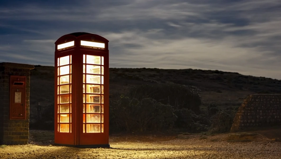 Illuminated red British telephone box standing alone in a rocky desert landscape at dusk.