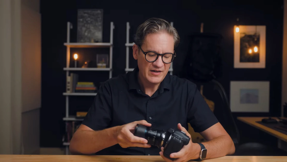 Man in black polo shirt examining a camera while seated at a wooden table in a studio setting.