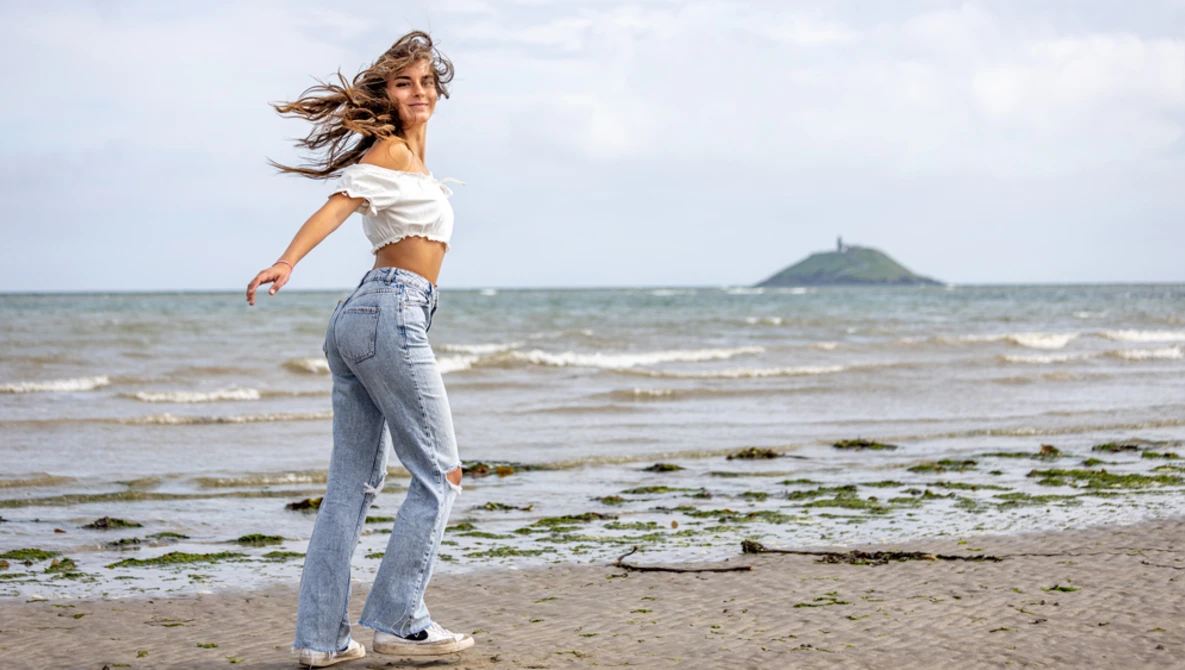 Woman in white off-shoulder top and jeans running on beach with ocean and distant peninsula in background.
