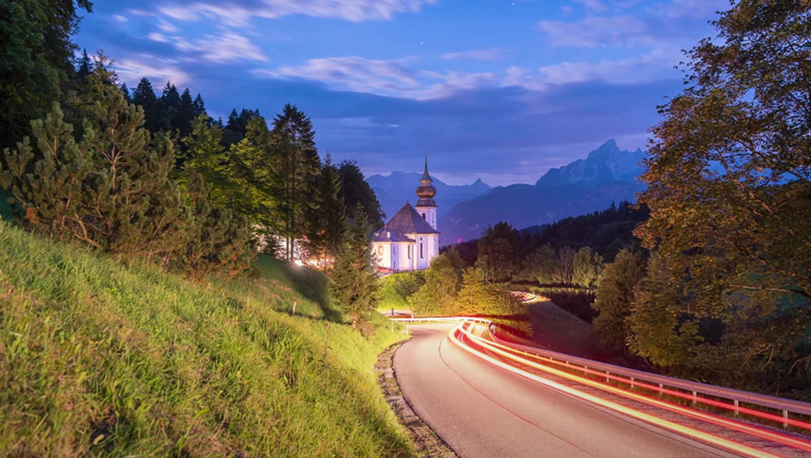Illuminated church with onion dome nestled in forested valley at twilight, with car light trails on curved road below.