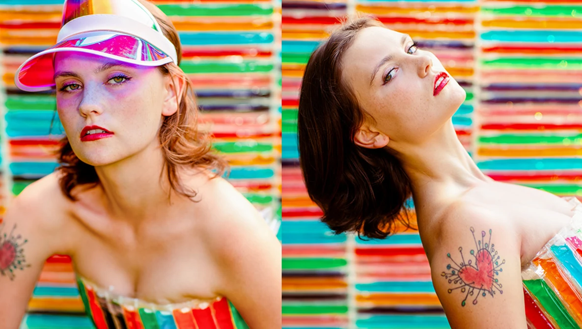 Two women posed against a vibrant multicolored striped background, wearing bold makeup and shoulder tattoos.