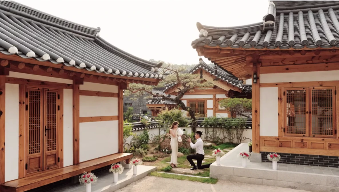 Traditional East Asian courtyard with red wooden buildings, curved tile roofs, and manicured garden with stone pathways.