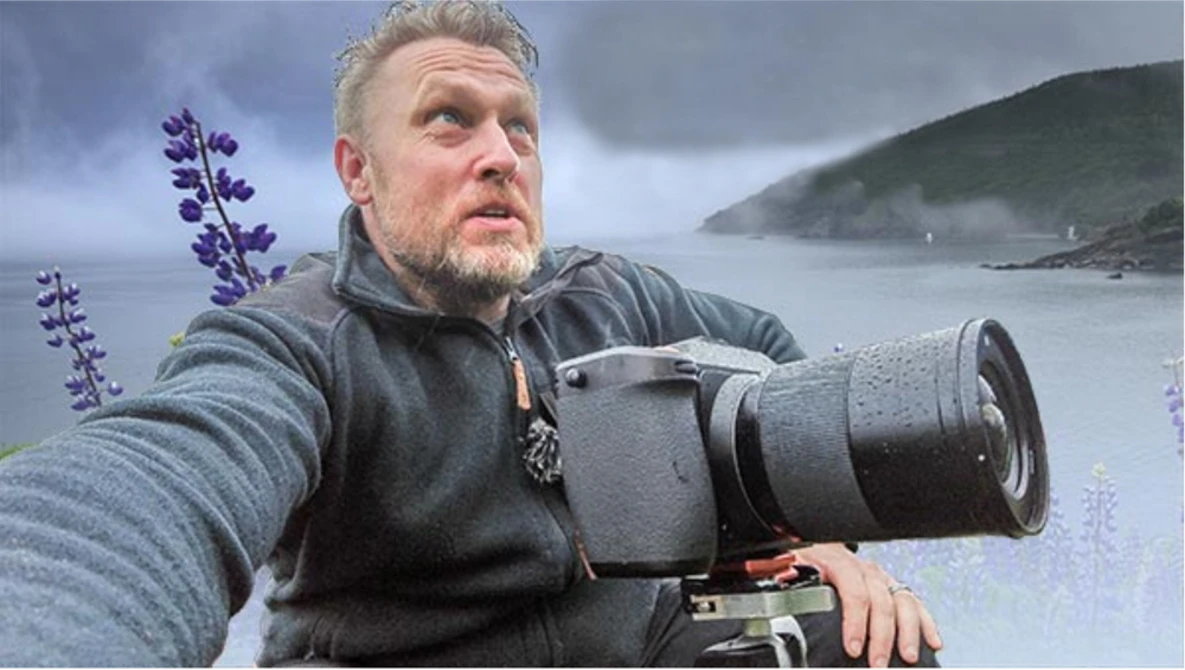 Photographer holding a telephoto lens while looking toward a coastal landscape with purple lupine flowers in foreground.