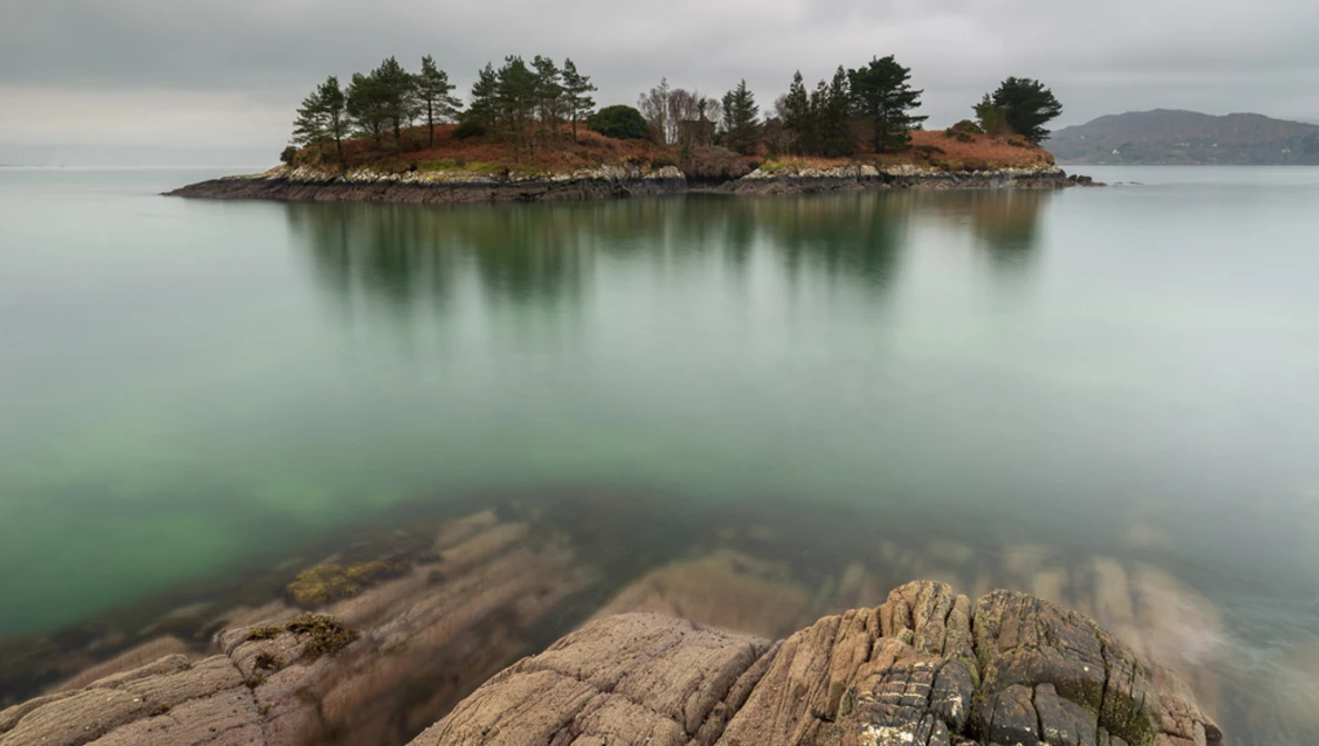 Rocky foreground with small forested island and its reflection in calm green water under overcast sky.