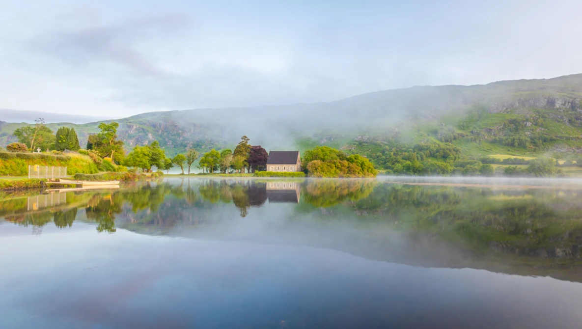 Misty river valley with stone building reflected in still water, surrounded by forested hills.