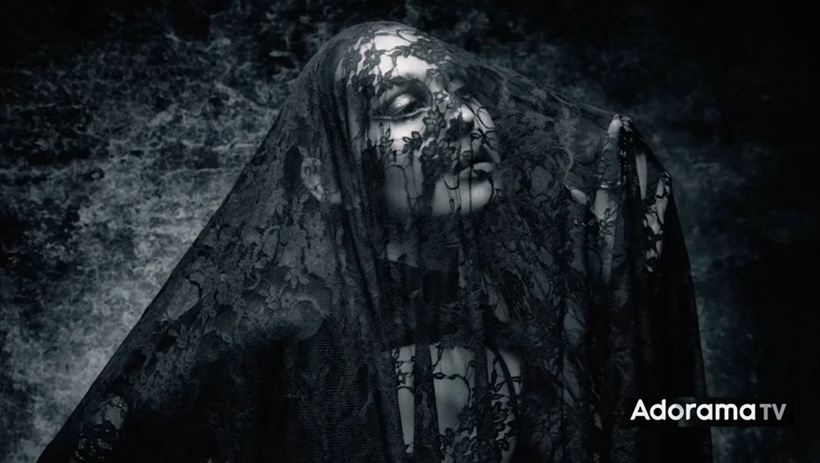 Figure draped in tattered black fabric against dark textured background with dramatic side lighting.