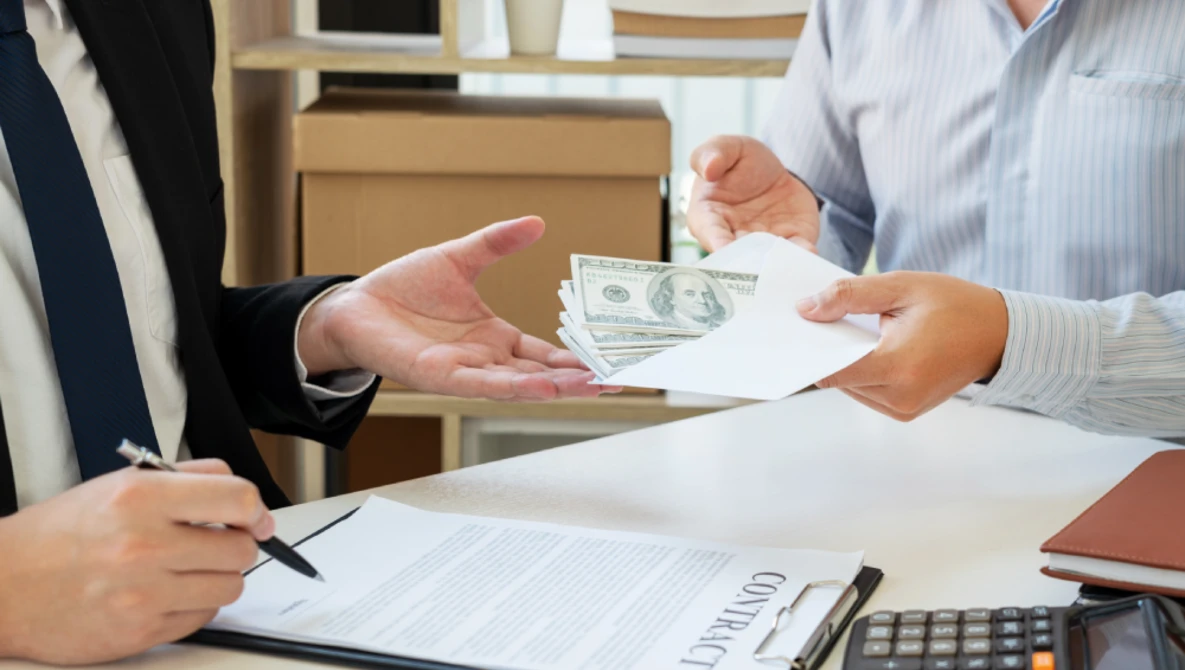 Two people exchange cash and documents at a desk during a business transaction.
