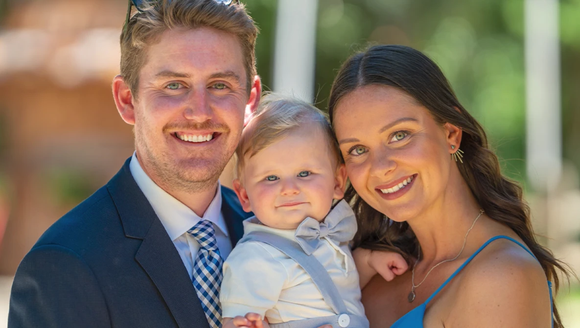 Smiling family of three poses together outdoors in natural sunlight.