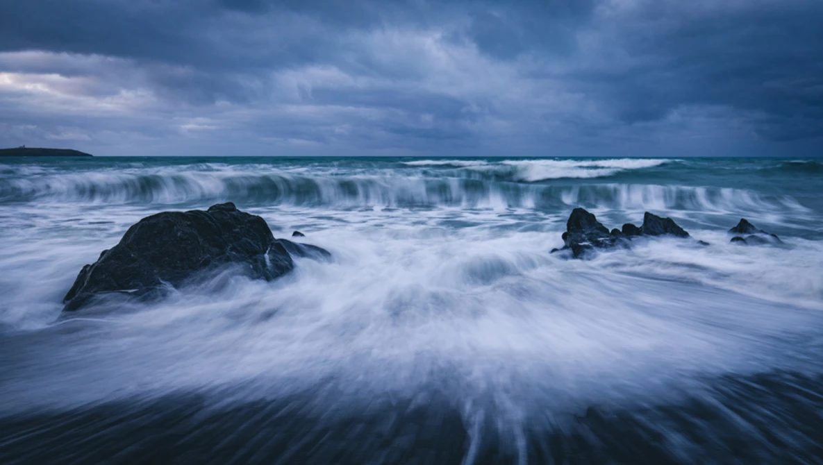 Waves crash over dark rocks on a beach under a dramatic stormy sky.
