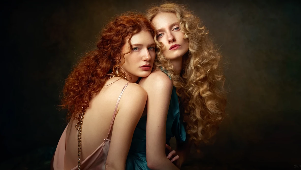 Two women with long curled hair posed closely together against a dark background.
