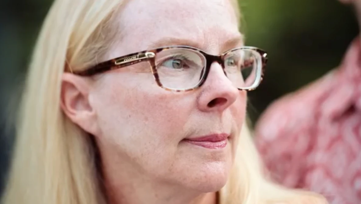 Woman wearing eyeglasses with blonde hair, looking thoughtfully to the side against a blurred green background.