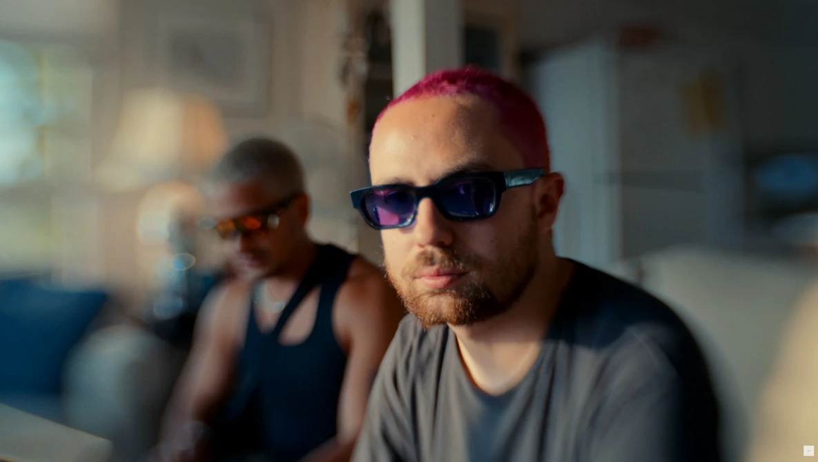 Man wearing blue-tinted sunglasses and gray t-shirt, shot indoors with natural window light creating warm highlights on his face.