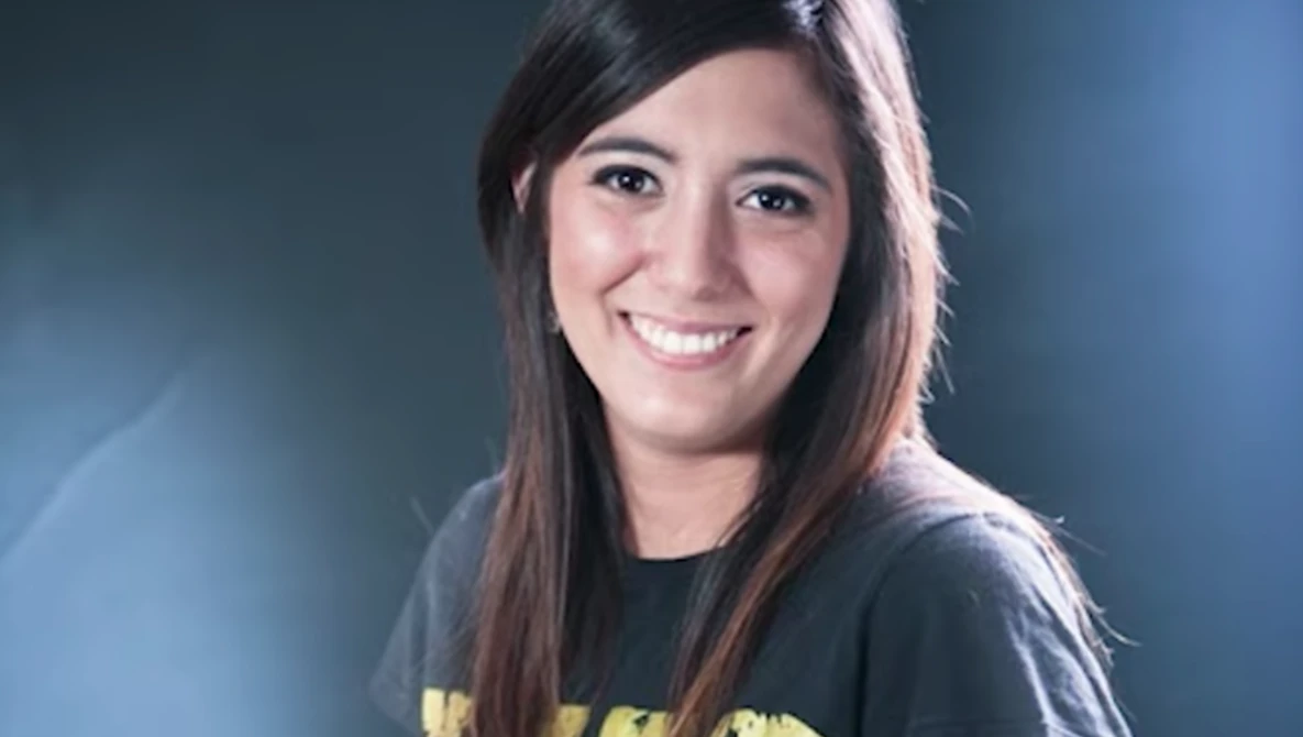 Young woman smiling at camera wearing dark graphic t-shirt against blue background.