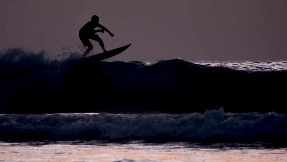 Silhouetted surfer riding a wave during golden hour with dramatic sky and ocean backdrop.