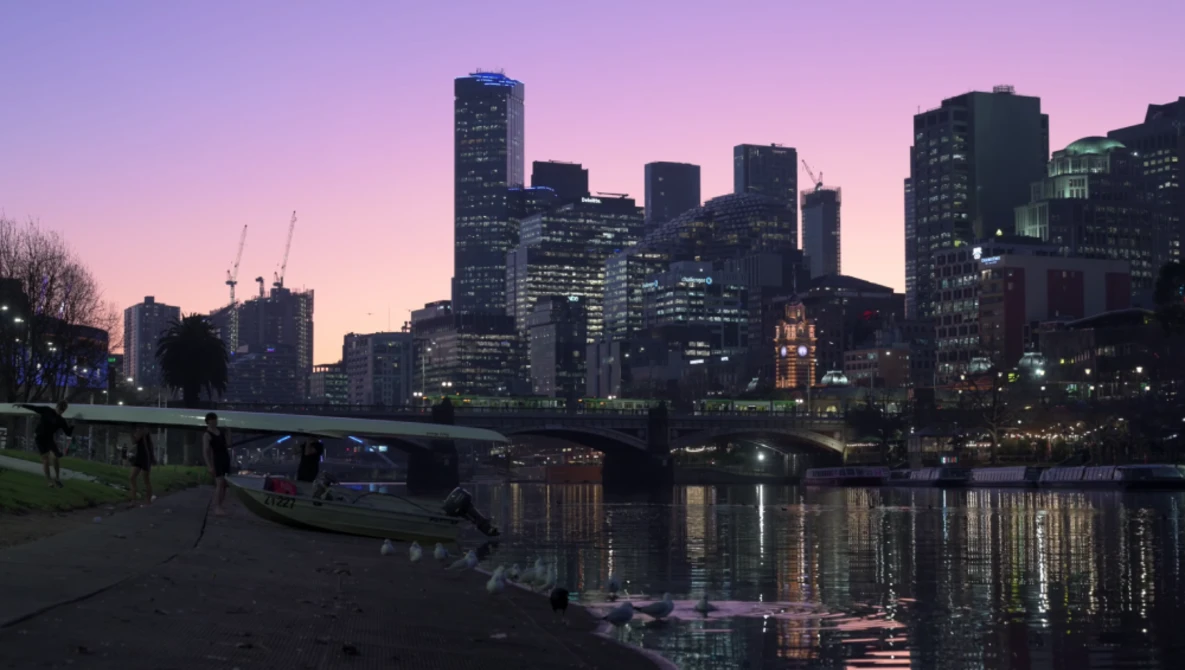 City skyline at twilight with illuminated skyscrapers reflected in still water below a bridge.
