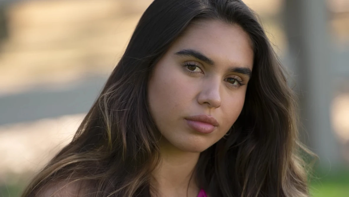 Young woman with long dark hair looking directly at camera with neutral expression.