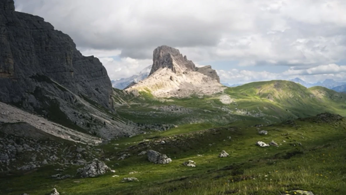Alpine valley with steep rocky peaks and lush green meadows under cloudy sky.