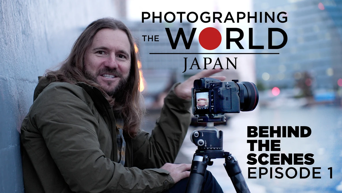 Photographer with long hair holding a medium format camera on tripod, smiling at camera in urban setting.