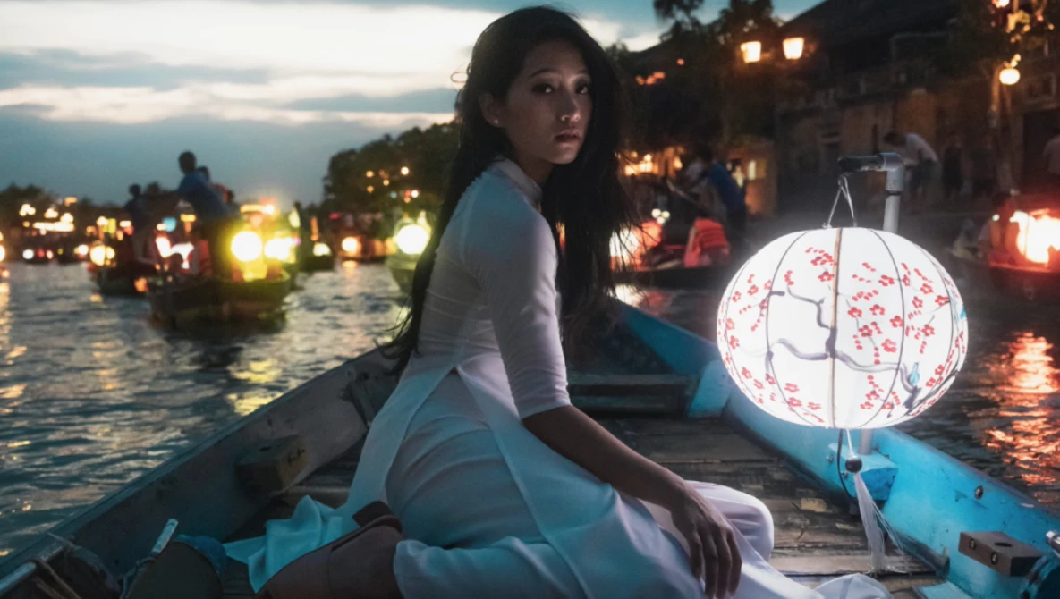 Woman in white clothing holding a decorative lantern in a boat at dusk among illuminated buildings.