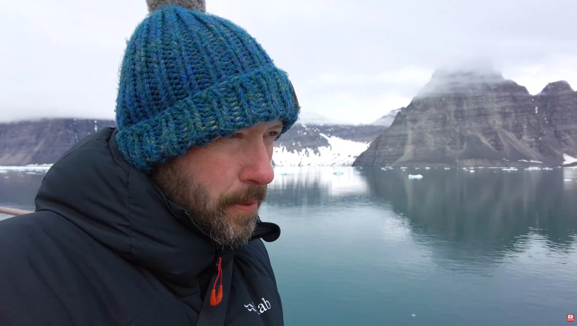 Man in blue knit beanie gazing toward snow-capped mountains across a glacial lake.