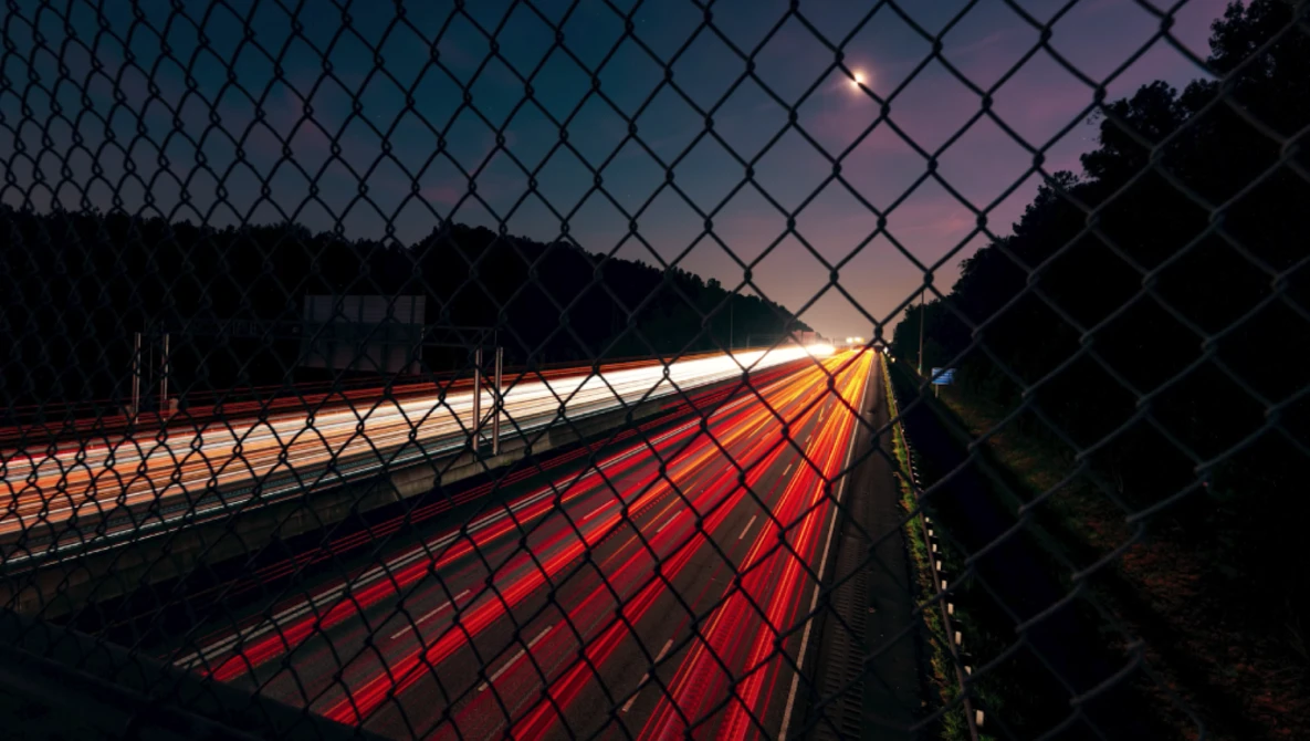 Highway at dusk viewed through chain-link fence, with red taillights creating light trails along the road.