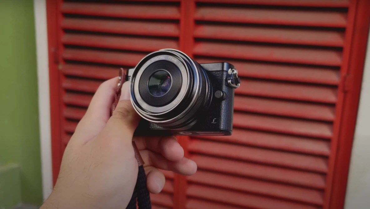 Hand holding a compact mirrorless camera with a wide-angle lens against a red louvered background.