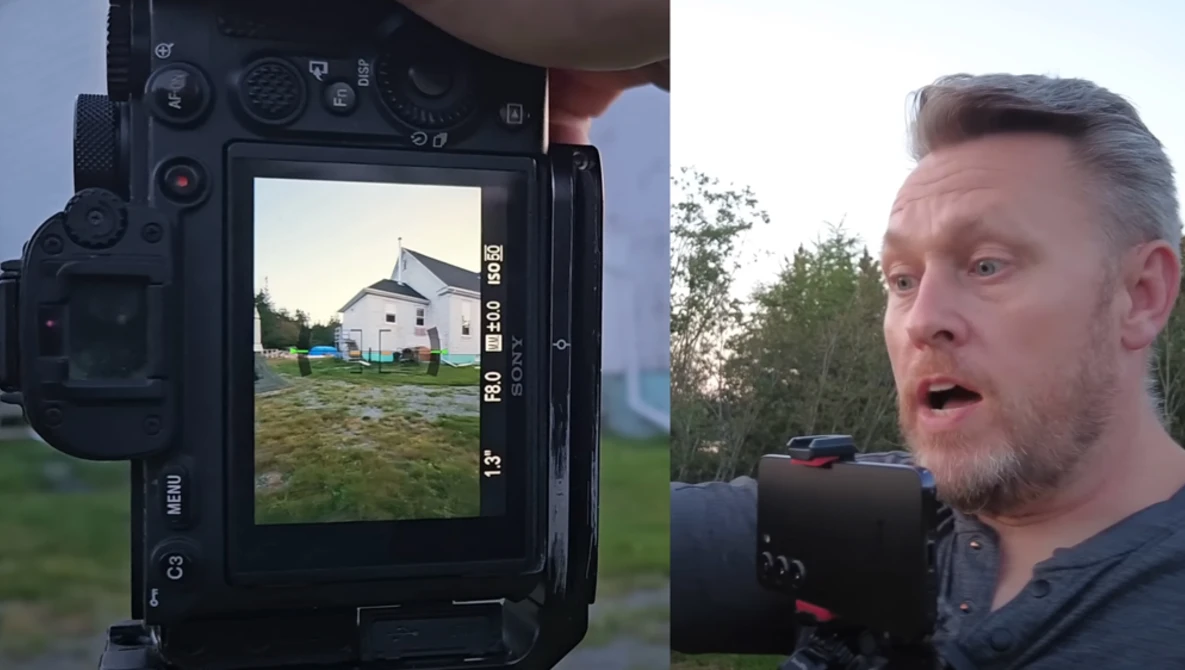 Photographer holding cinema camera while monitoring a residential property on the camera's rear display screen.