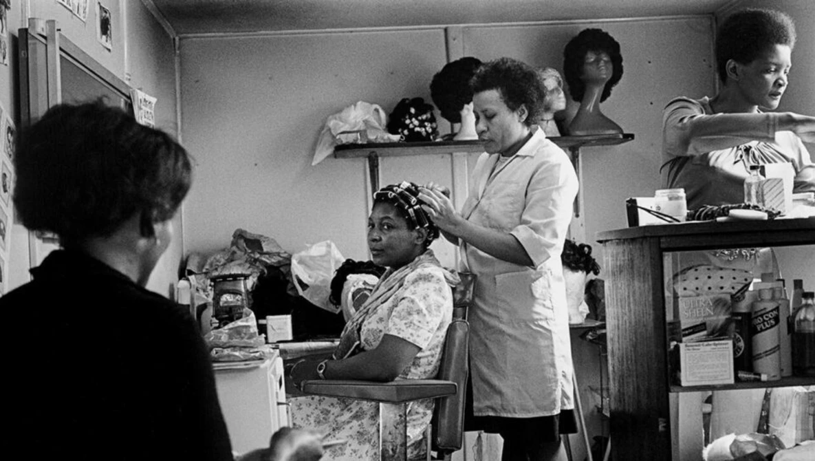 A woman having her hair styled by another woman in what appears to be a beauty salon or barbershop interior.
