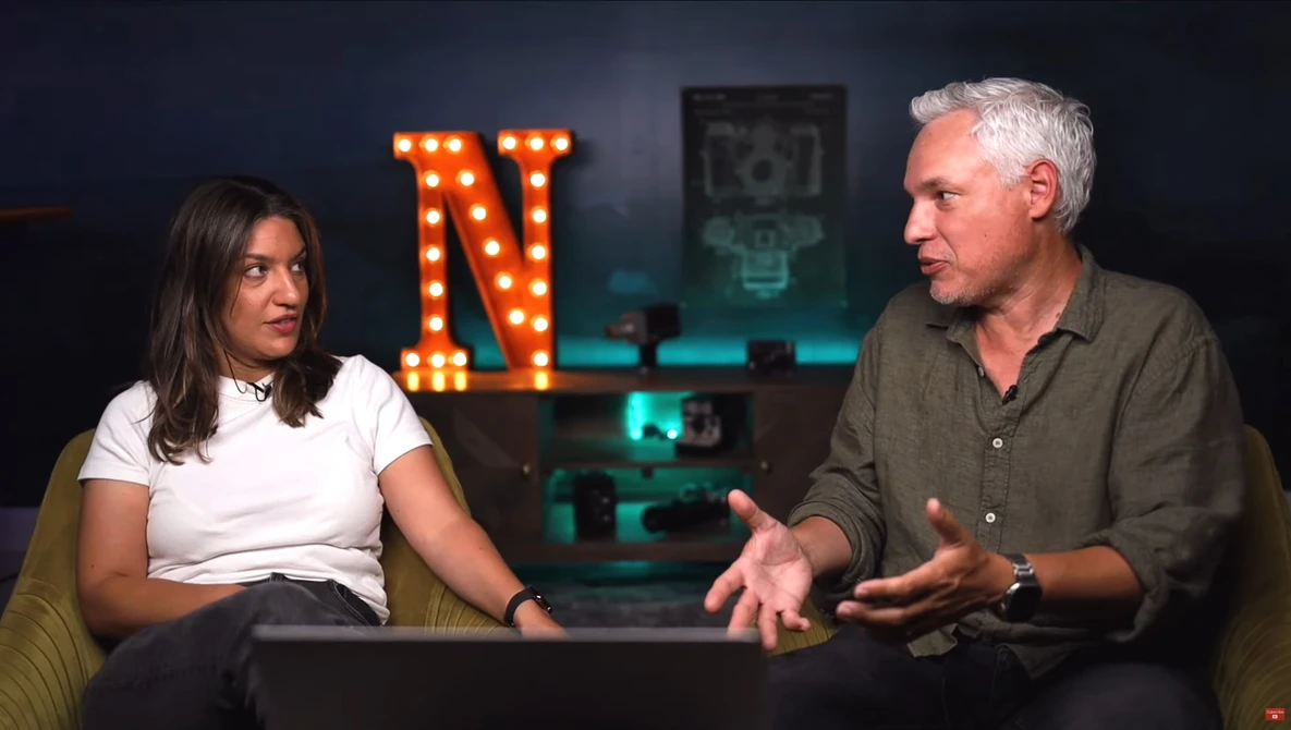 Two people in conversation during an interview or podcast recording set with neon signage in the background.