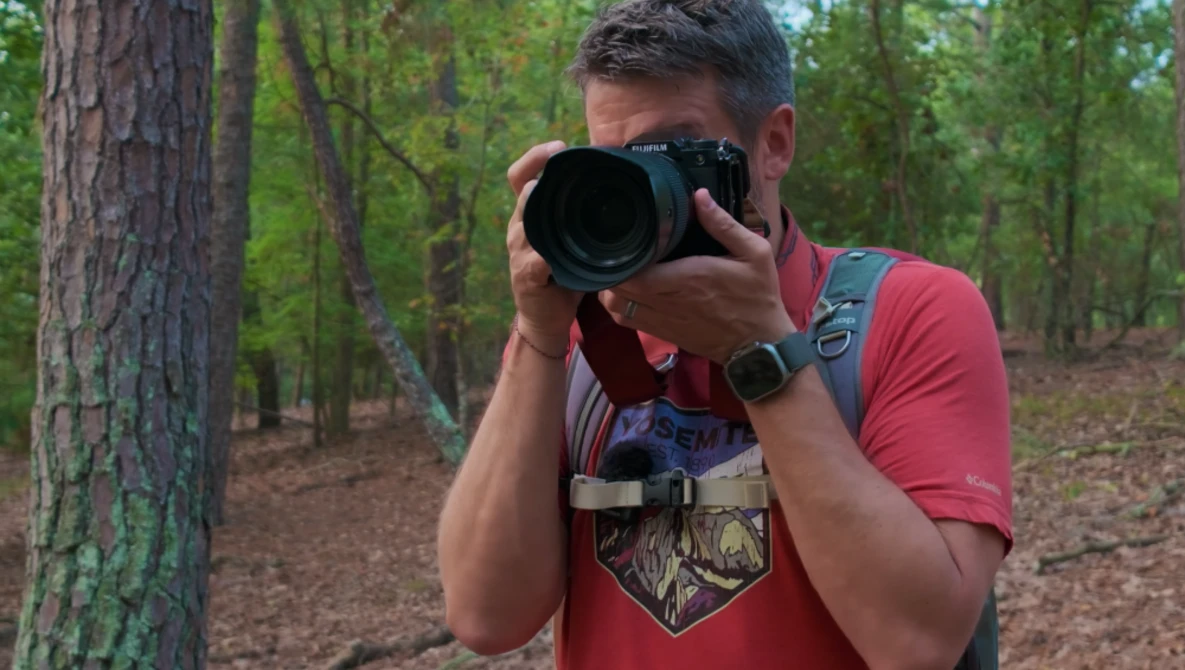Photographer holding a telephoto lens camera in a forest setting, wearing a red shirt and backpack.