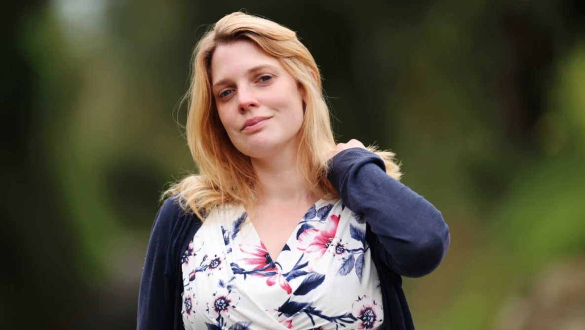 Woman with blonde hair wearing a floral print blouse and navy cardigan, photographed outdoors with a soft, blurred green background.