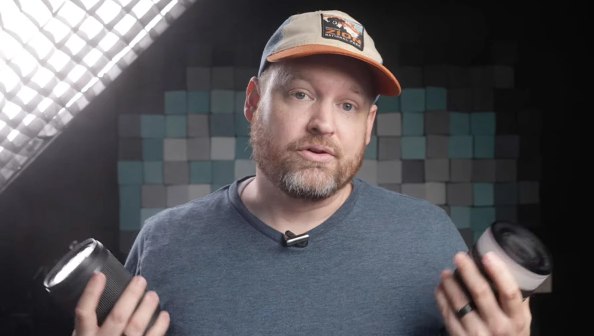 Man in baseball cap holding two cameras while speaking to camera in studio setting.