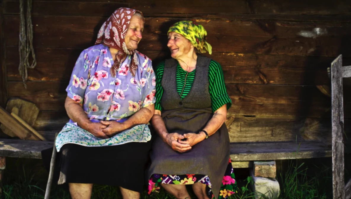 Two women seated on a wooden bench against a dark wood wall, smiling at each other in warm conversation.