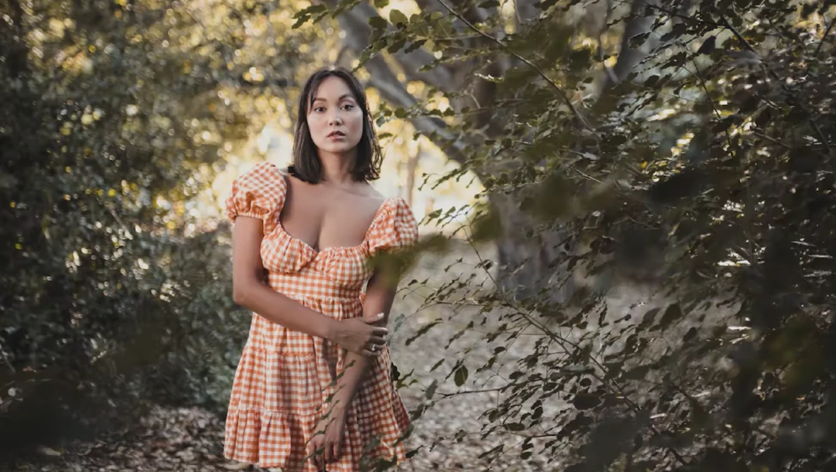 Woman in orange gingham dress posing among green foliage and tree trunks.