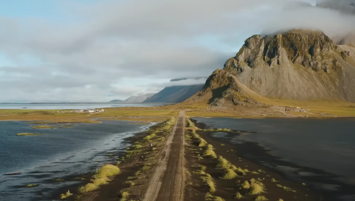 Gravel road stretching toward a distinctive peaked mountain across black sand plains with moss-covered terrain.
