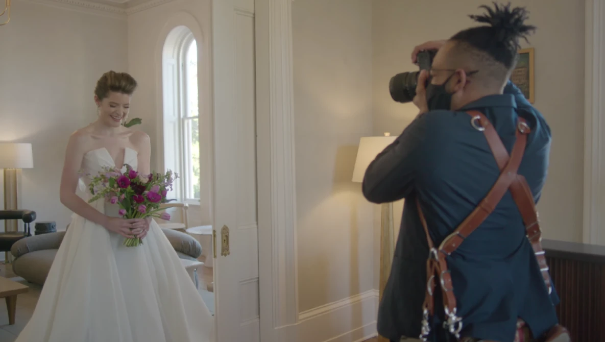 Photographer with camera on shoulder captures bride in white gown holding purple bouquet in elegant hallway.
