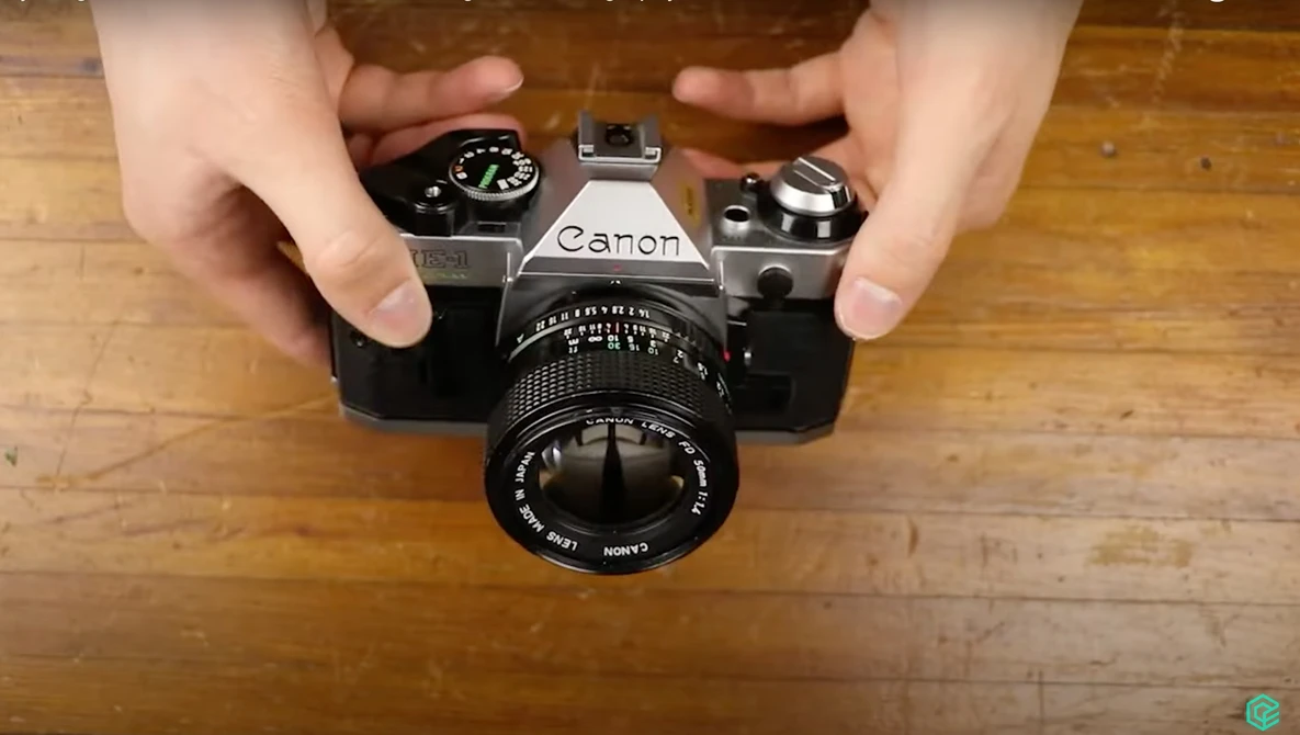 Hands holding a vintage Canon SLR camera with manual lens on wooden surface.